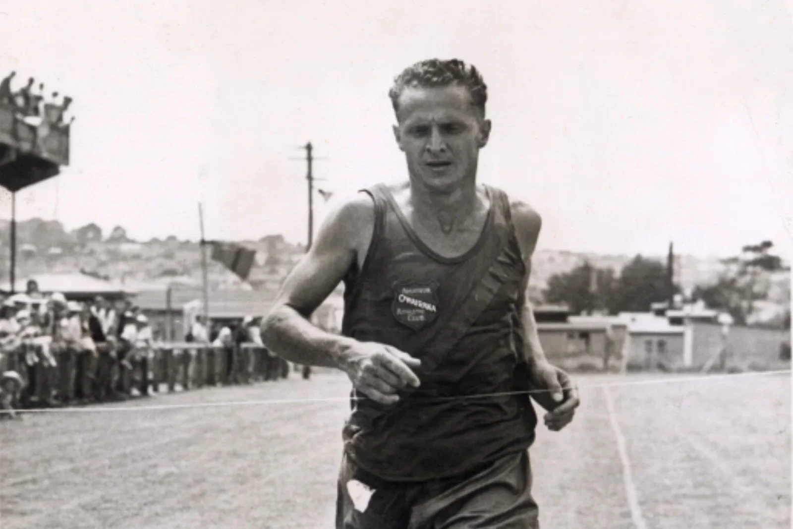 Black-and-white photo of a young Arthur Lydiard breaking the finishing tape in an Owairaka Athletic Club vest